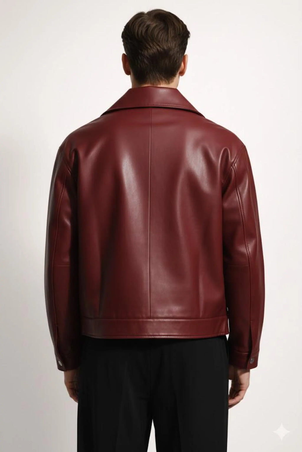 A man viewed from the back wearing a glossy burgundy leather jacket and black trousers, standing against a plain light gray studio background.