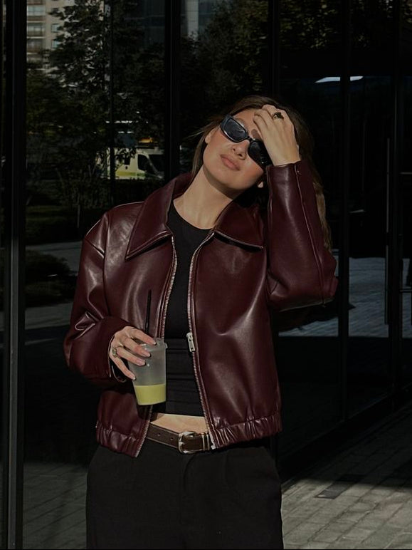 A woman wearing a dark burgundy leather jacket, black top, and black pants stands in front of a modern glass building at night, holding a drink.