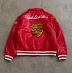 A red Porsche leather jacket shown from the back, featuring a large embroidered Porsche crest on the center back, white cursive text reading 'Aime Leon Dore' across the shoulders, and white and red striped ribbed cuffs and hem, laid flat on a gray textured surface.