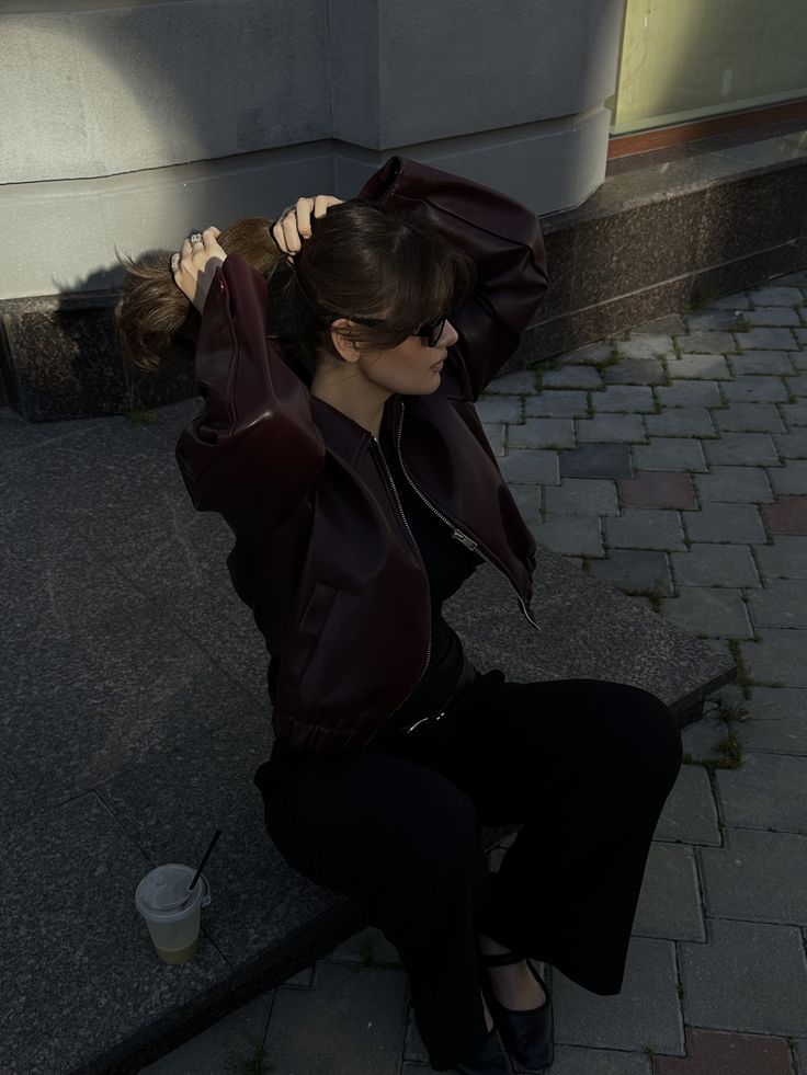 A woman sitting on a stone curb outdoors, wearing a Maroon Faux Leather Jacket jacket, black pants, and black shoes. She is adjusting her hair with both hands, wearing dark sunglasses.