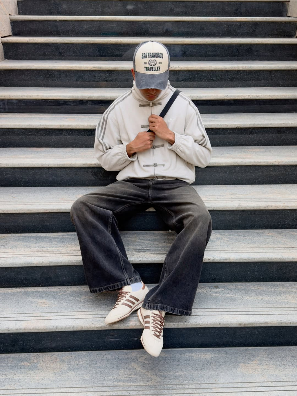 A person sitting on outdoor concrete stairs wearing a light gray zip-up Cny jacket with three stripes on the sleeves, dark wide-leg jeans, white sneakers, and a gray and white baseball cap.